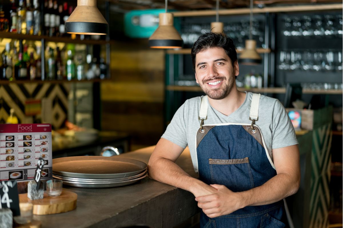 a UK business owner thinks about his electricity supply as he leans against a counter in a restaurant wearing a blue apron