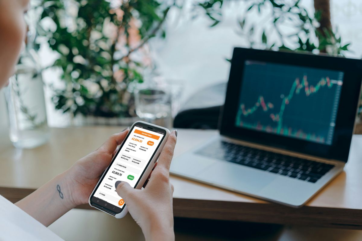 Someone using a mobile phone to check the latest business gas prices. a laptop in the background shows a line graph going up and down.