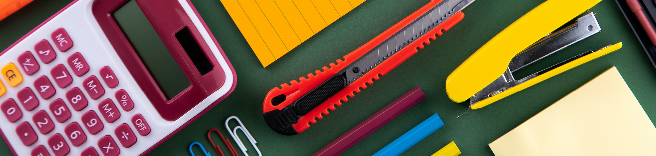 different school stationery supplies on a dark green background, including a pink calculator, yellow stapler and sticky notes