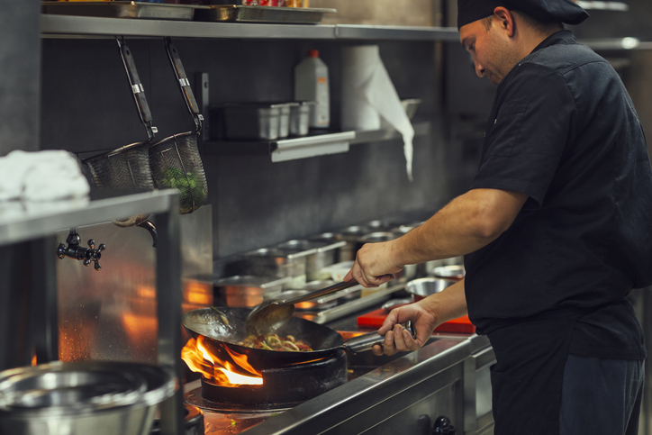 Chef preparing asian food in restaurant kitchen. There are flames under the pan from a gas hob. The kitchen is modern. The chef is thinking about switching his business gas supplier.