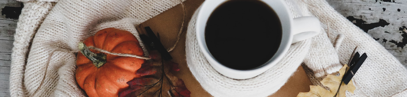 A mug of black coffee sat on top of a notebook in haloween marketing style, with a small pumpkin and cosy winter scarf