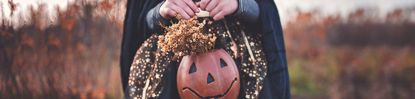 Close up of a childs hands holding a pumpkin bucket filled with flowers. She is wearing a halloween costume.