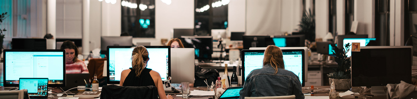 Office Energy Saving Tips - An image of multiple people sat at desks in an atmospherically lit office. They are using desktop computers with their back to the camera.