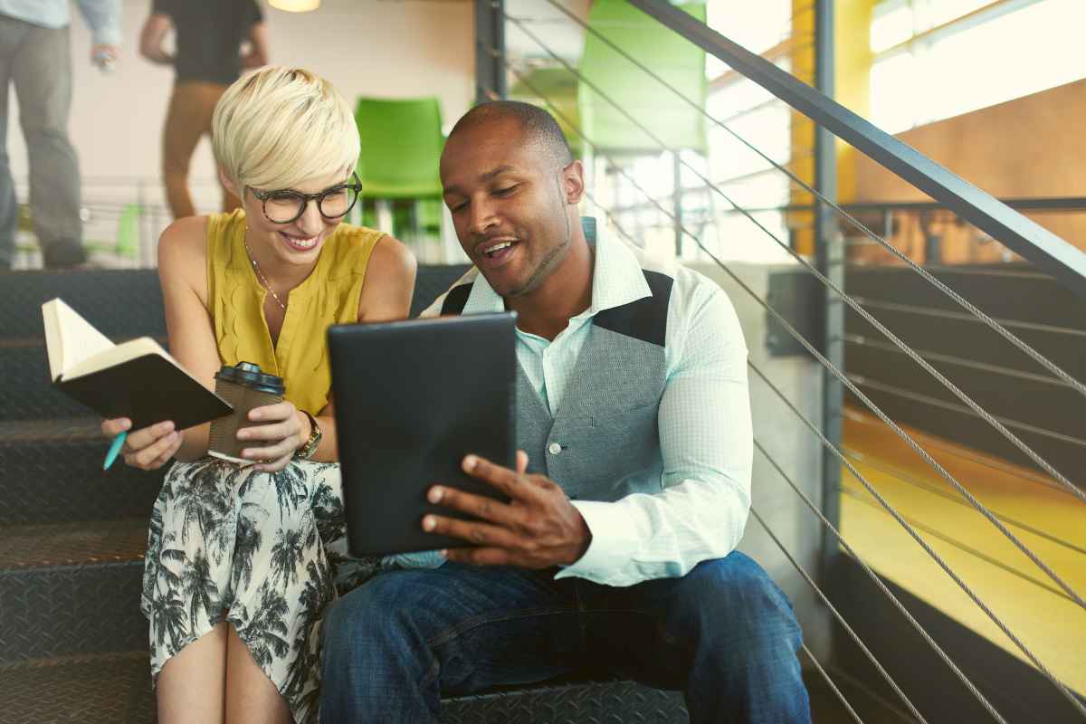 Two collegues reviewing their business checklist whilst sat on the stairs. They both look happy. The female colleague is on the left with a notebook, and her male colleague holds an ipad.