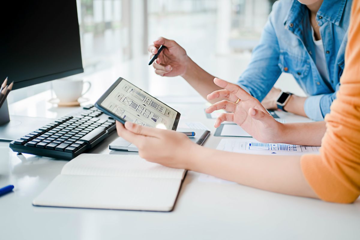 Cropped closeup of two business people creating a business website together. They are holding an ipad with UX sketches in a notebook underneath.