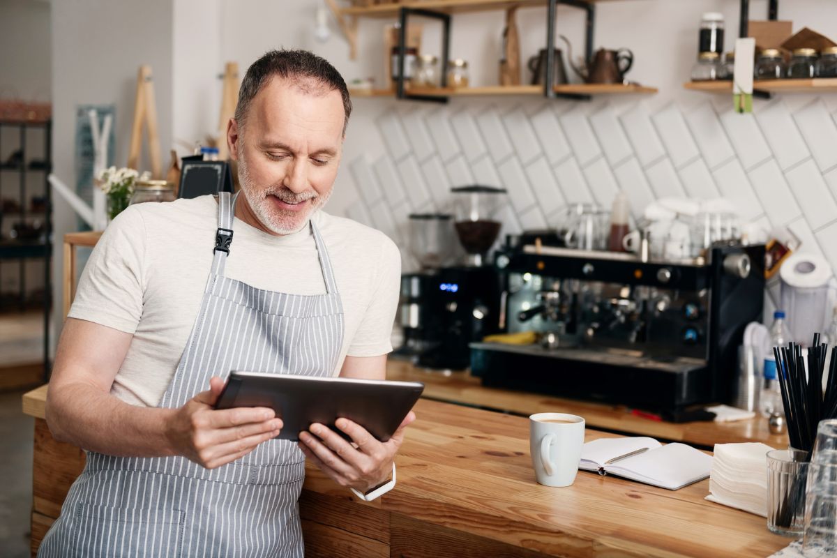 A man in a white tshirt and blue striped apron compare suppliers on a tablet whilst considering the best time to switch business energy. he is leaning on a wooden counter in a cafe.