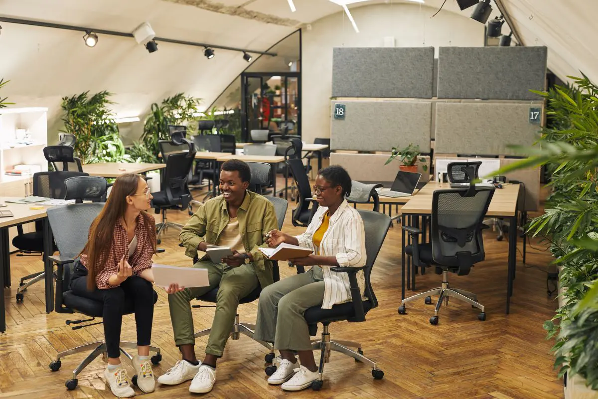 three colleagues in a modern office discussing the benefits of renewable energy for businesses. The office has a wooden floor and lots of greenery around it.