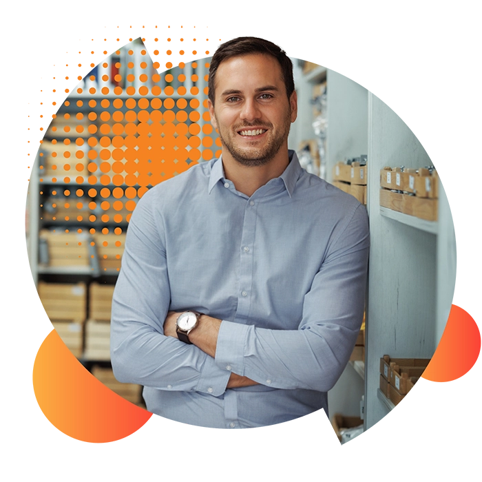 A man with arms folded, dressed in a pale blue shirt, leans against the shelves in his business after choosing his small business energy supplier